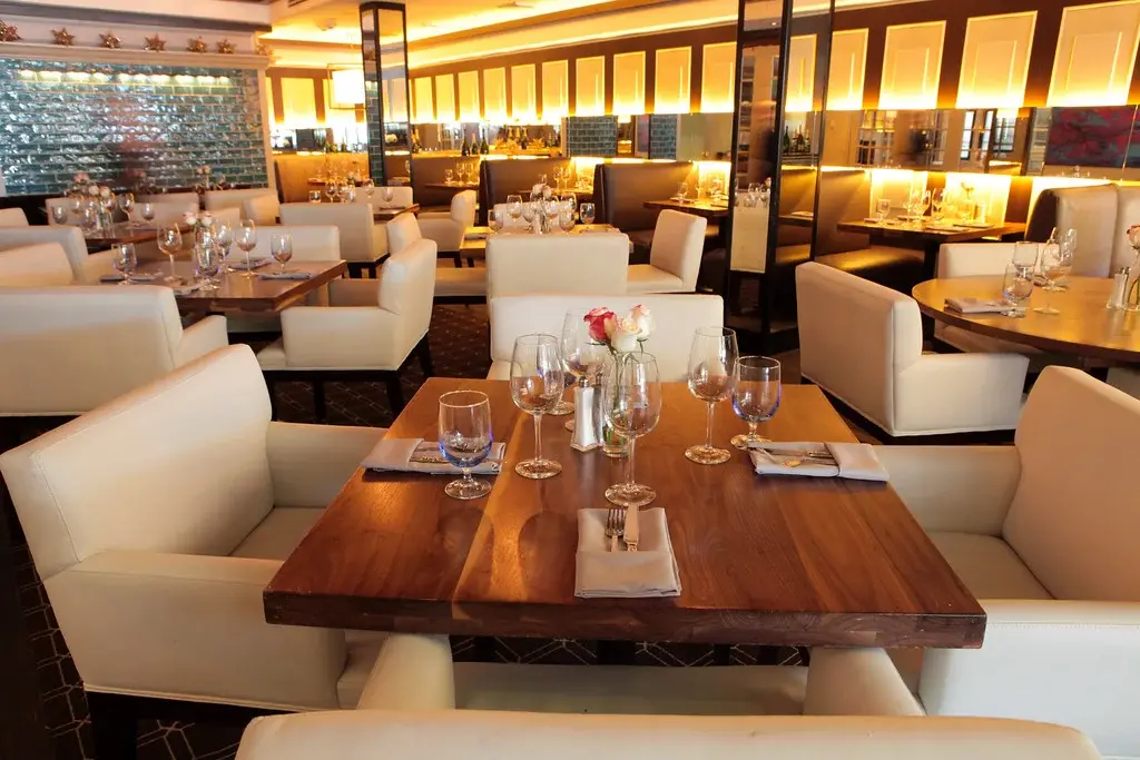 Modern upscale restaurant interior with white upholstered seating, wooden tables set with wine glasses and flowers, and backlit window panels creating warm ambient lighting.